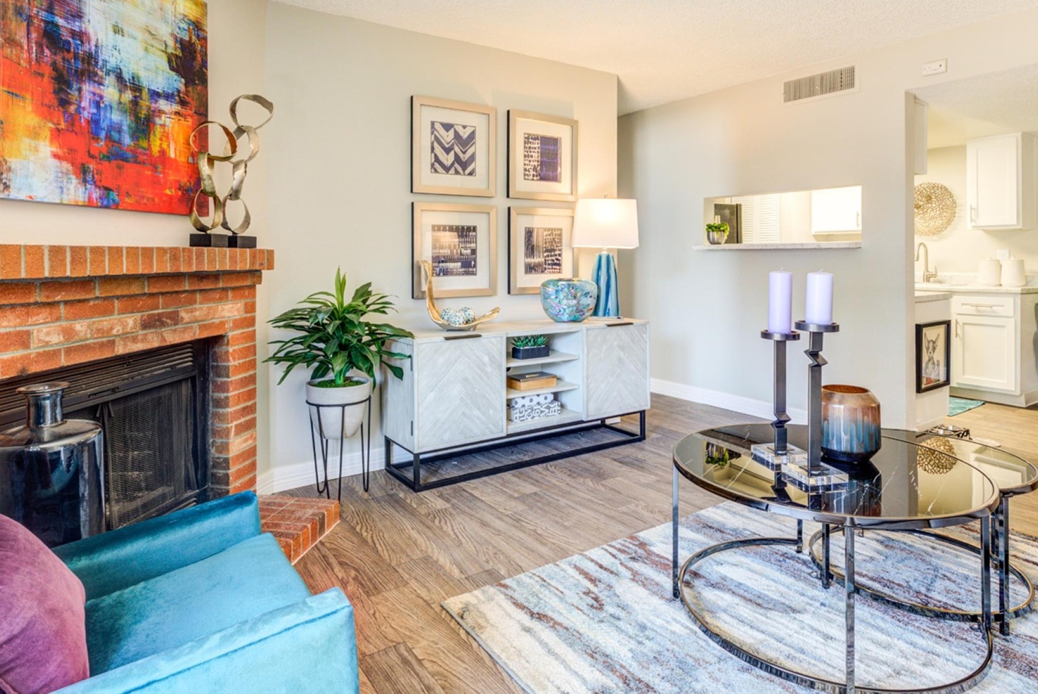 A stylish and contemporary living room at Santana Ridge featuring a cozy brick fireplace with vibrant abstract artwork above it. The room is furnished with a teal accent chair, a modern white console table adorned with decorative pieces, and a sleek glass coffee table with candles. The open-concept design leads to a dining area and kitchen with white cabinetry, creating a bright and inviting space. Warm wood flooring and indoor plants add a touch of comfort and elegance.