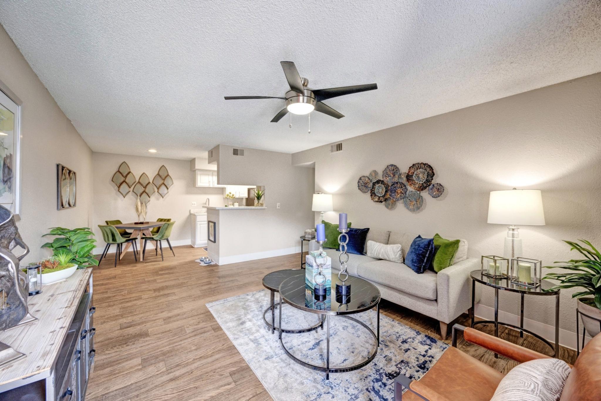 A modern open-concept living space at Skyline Apartments featuring a stylish living room, dining area, and kitchen. The living room is furnished with a gray sofa adorned with blue and green accent pillows, a round glass coffee table, and a leather accent chair. Decorative wall art, table lamps, and lush indoor plants enhance the ambiance. The dining area includes a wooden table with four green velvet chairs, while the kitchen features white cabinetry and a pass-through counter, creating a seamless and invit