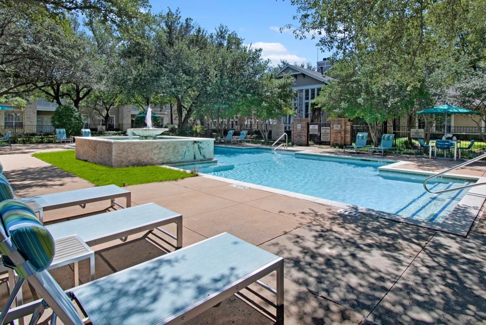 A luxurious outdoor pool area in a residential community, surrounded by mature trees and lush landscaping. The pool features a built-in water fountain, lounge chairs with blue and green striped pillows, and patio tables with teal umbrellas. A modern clubhouse with large windows is visible in the background, adding to the relaxing, resort-like atmosphere. ​​