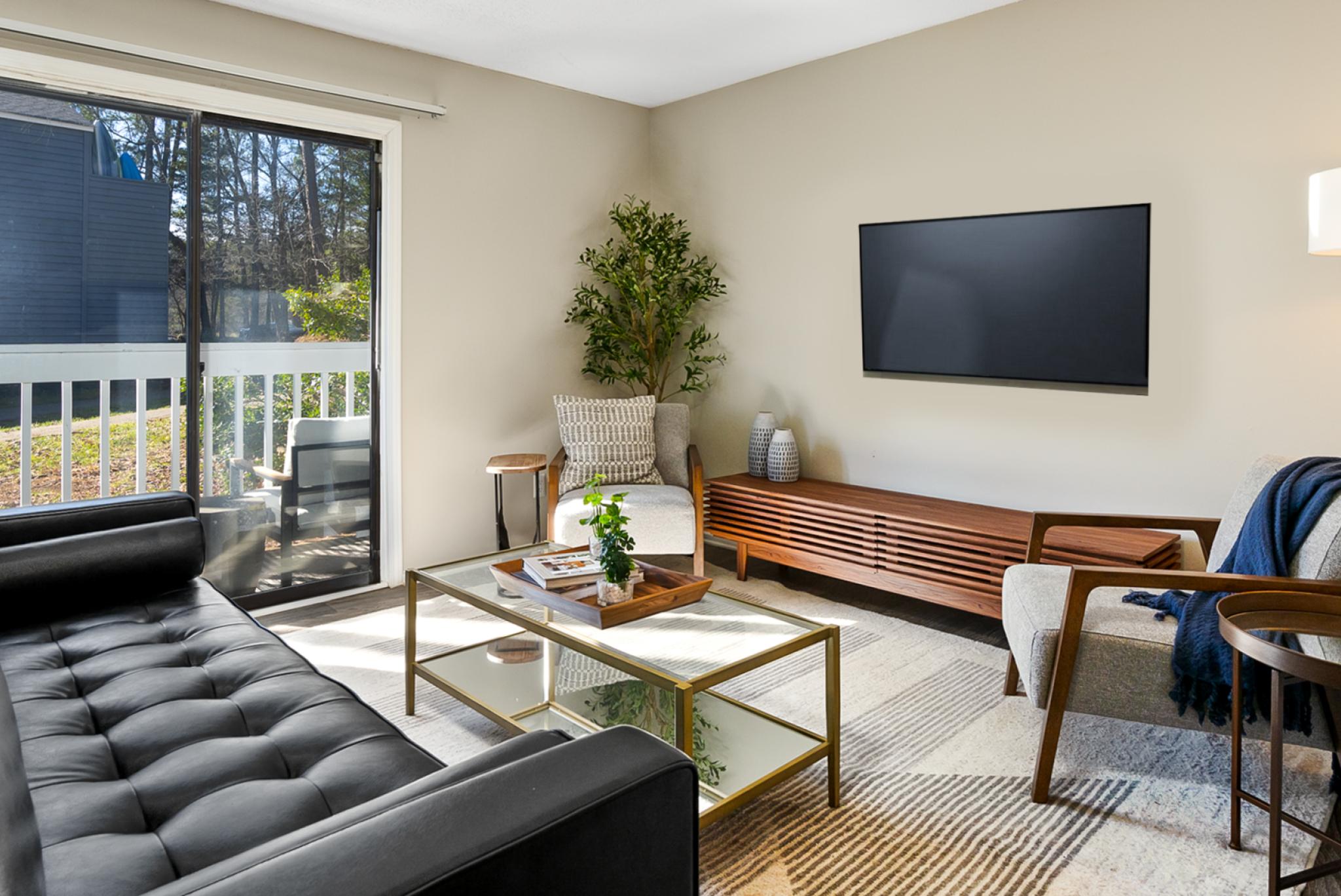 Modern living room at The Forest Apartments featuring a black leather sofa, a glass coffee table with a gold frame, and a cozy armchair with a throw blanket. A large sliding glass door leads to a private balcony with outdoor seating, allowing natural light to fill the space. The room is decorated with neutral tones, a sleek media console, and stylish accents, creating a warm and inviting atmosphere.