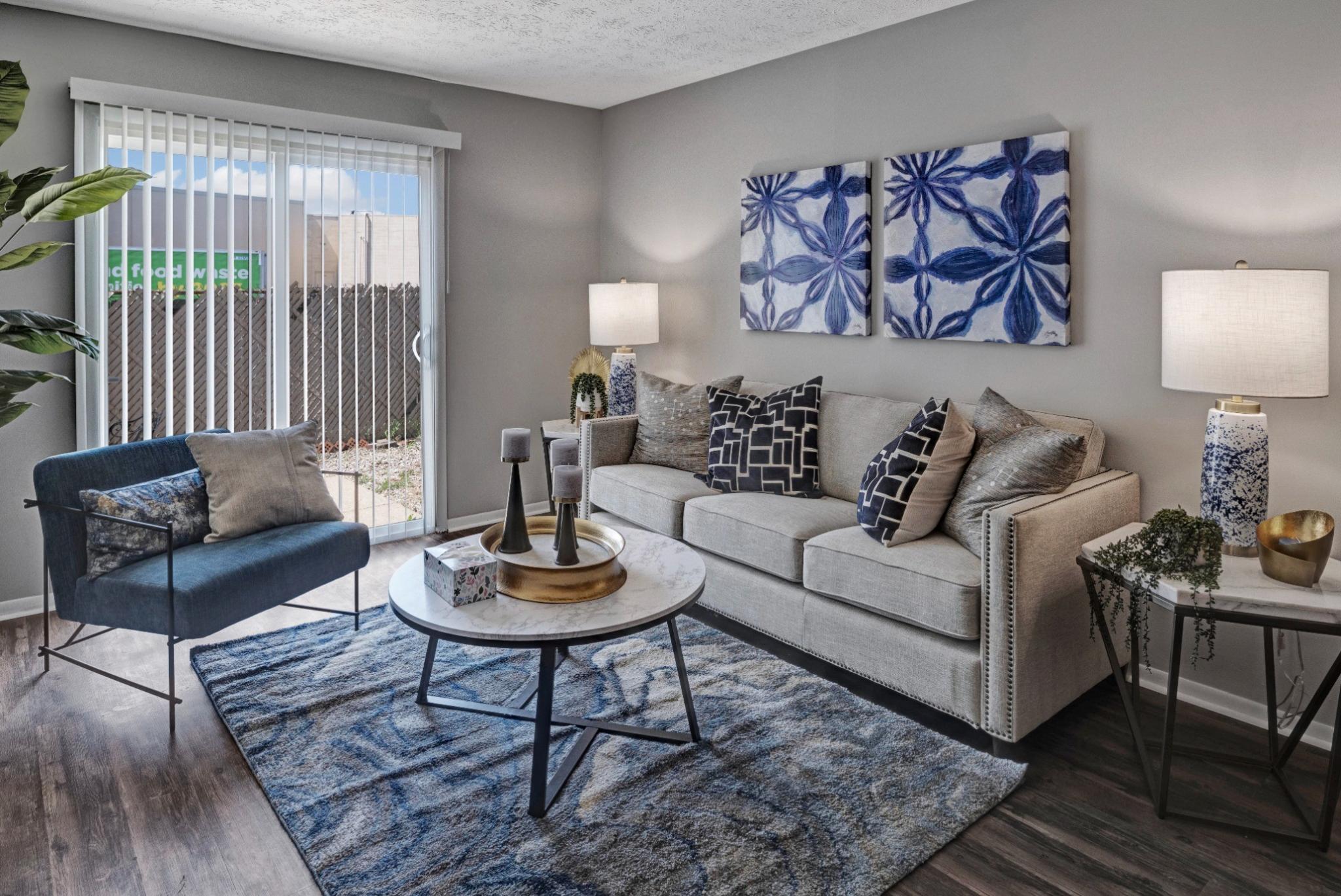 A stylish living room at Wilson Plaza featuring a beige fabric sofa adorned with decorative throw pillows, a navy blue accent chair, and a marble coffee table with a gold tray centerpiece. The space is illuminated by modern table lamps, and the decor includes blue abstract wall art and a patterned area rug. A sliding glass door with vertical blinds leads to an outdoor patio area, bringing in natural light. ​​