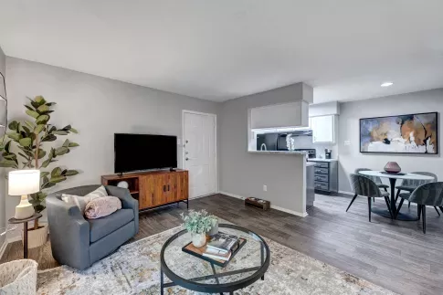 A modern living room with gray walls, wood flooring, a cozy seating area, a TV console, and an open view into a compact kitchen and dining space.