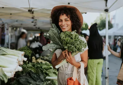 A woman smiles while holding fresh greens at a vibrant outdoor farmers' market.