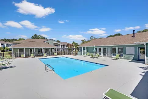 An outdoor swimming pool surrounded by green lounge chairs and a light blue clubhouse under a bright blue sky with fluffy white clouds.