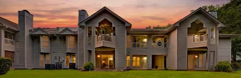 A two-story residential building with balconies, surrounded by greenery, set against a vibrant sunset sky.