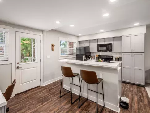 A modern kitchen with gray cabinetry, a marble countertop breakfast bar, and bar stools, complemented by hardwood floors and ample natural light from large windows.
