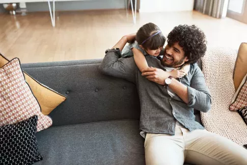 A father and daughter share a joyful moment, hugging and laughing on a modern grey sofa in a bright living room.