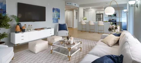 A bright and modern living room with white and gold decor, a mounted TV, and a view into an open-concept kitchen with a bar area.