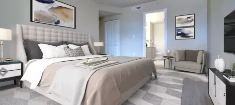 A stylish bedroom with a plush bed, elegant neutral tones, a cozy armchair, and a bathroom visible through an open door.