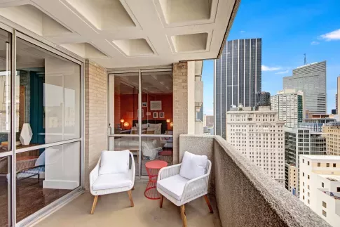 A cozy apartment balcony with white chairs and a red side table, overlooking a vibrant cityscape.