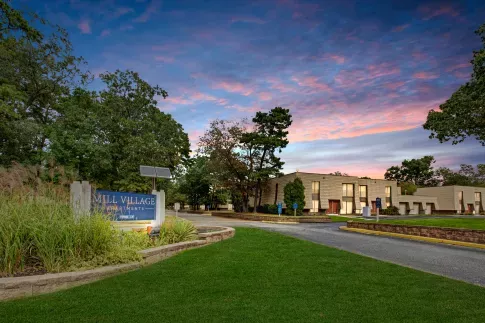 A welcoming entrance to Mill Village Apartments with lush greenery and a vibrant sunset sky.