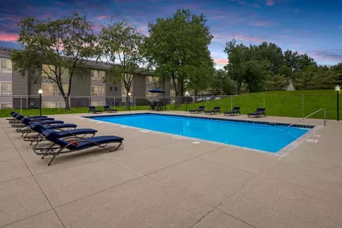 Outdoor swimming pool surrounded by lounge chairs, with a sunset sky and a grassy area in the background.