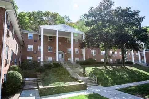 A charming brick apartment building at The Manors Apartments, surrounded by lush greenery and featuring classic white columns at the entrance, offering a welcoming and serene living environment.