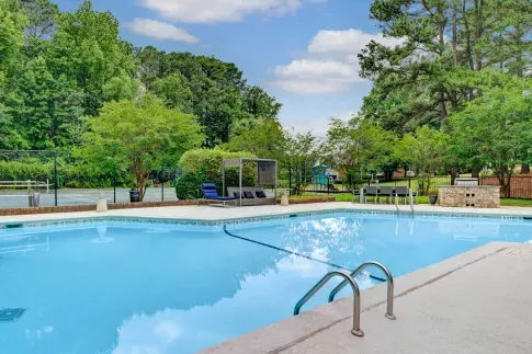 A serene outdoor pool area at Aria North Hills, surrounded by lush greenery and tall trees. The pool features stainless steel handrails, lounge chairs, and a shaded cabana area for relaxation. A tennis court and a children's playground can be seen in the background, offering a variety of recreational amenities. ​​