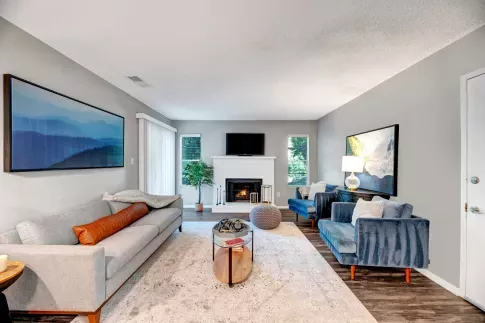 A beautifully designed living room at Aria North Hills featuring a neutral color scheme with blue accent chairs and a cozy fireplace. The room is furnished with a sleek gray sofa, a modern glass coffee table, and elegant artwork on the walls. Large windows and sliding doors allow natural light to brighten the space.