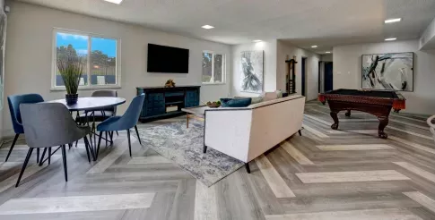 A stylish community lounge with a modern design, featuring a light beige sofa with blue accent pillows, a dark wood pool table with orange tassels, and a round dining table with blue chairs. The room includes a sleek entertainment center with a mounted flat-screen TV, large windows allowing natural light, and abstract art pieces on the walls. The flooring is arranged in a chevron pattern with shades of gray and beige. ​​