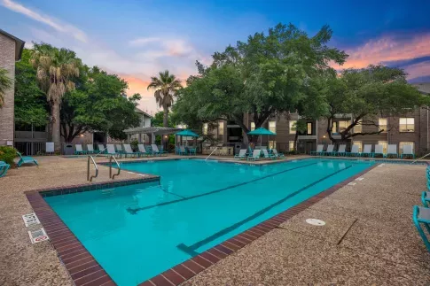 Resort-style swimming pool surrounded by tall palm trees, mature shade trees, and apartment buildings, with plenty of teal lounge chairs and umbrella-covered seating areas on a textured pool deck at sunset.
