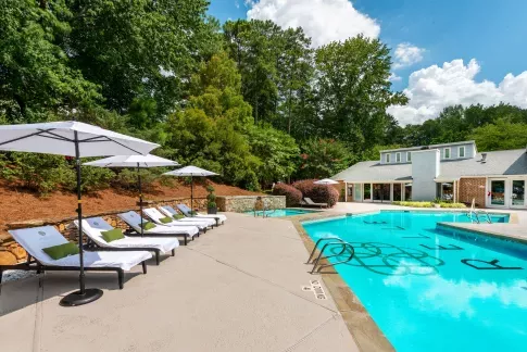Inviting outdoor pool area with cushioned lounge chairs and white umbrellas, surrounded by lush landscaping and a clubhouse with large windows, offering a tranquil and stylish retreat.