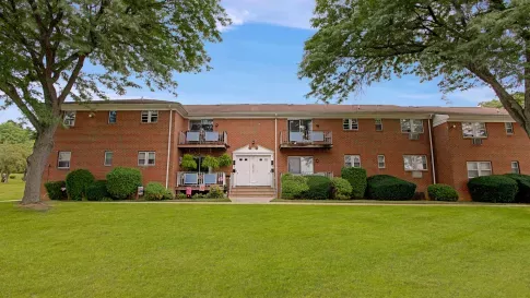 Charming brick apartment building with balconies and well-manicured landscaping, set against a backdrop of mature trees and a wide, green lawn under a clear blue sky.