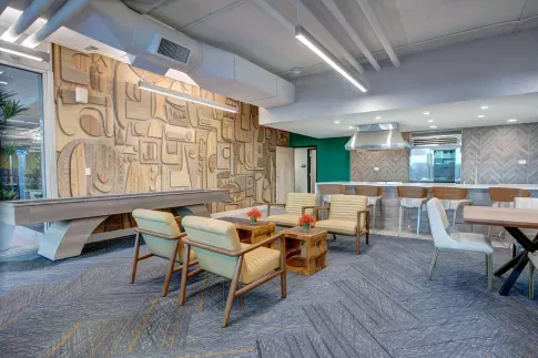 Stylish resident lounge with modern decor, featuring a shuffleboard table, mid-century seating, geometric wood accent wall, and a sleek demonstration kitchen with bar seating and stainless steel appliances.