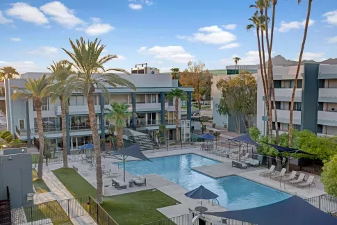 Resort-style pool area surrounded by modern apartment buildings and tall palm trees, featuring two swimming pools, shaded lounge areas with umbrellas and sails, and scenic mountain views in the background.