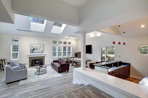 A spacious and modern clubhouse at Chandlers Bay featuring an open-concept design with high ceilings and skylights. The room is furnished with a brown leather sofa, gray armchairs, and a contemporary fireplace with a white mantel. A stylish wooden pool table is positioned near a wall-mounted TV, and a built-in bar area with granite countertops and red pendant lights adds to the upscale ambiance. Large windows and glass doors allow natural light to fill the space, offering views of the outdoor greenery. ​​