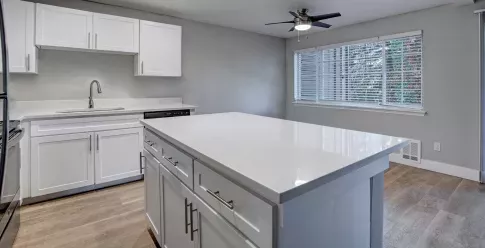 A modern kitchen at Chandlers Bay featuring sleek white cabinetry with silver hardware, quartz countertops, and a spacious center island with ample storage. The kitchen is equipped with a stainless steel sink and a high-arc faucet. Large windows with blinds allow natural light to brighten the space, and a ceiling fan with a built-in light fixture adds functionality. The open floor plan connects seamlessly to the adjacent living area, creating a contemporary and inviting atmosphere. ​​