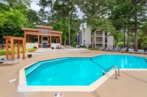 Hexagonal outdoor swimming pool surrounded by lounge chairs, with a modern wooden pergola and grill area set against a lush wooded backdrop and apartment buildings.