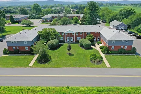 Aerial view of a well-maintained brick apartment building with neatly trimmed landscaping, surrounded by lush greenery and distant rolling hills.
