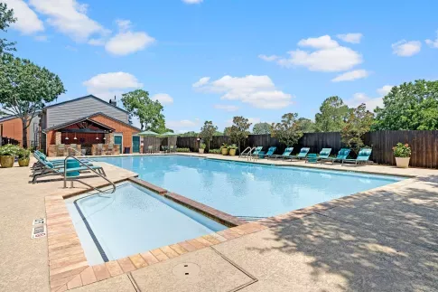 Resort-style swimming pool with poolside lounge chairs, shaded seating area, and a red brick clubhouse under a clear blue sky.