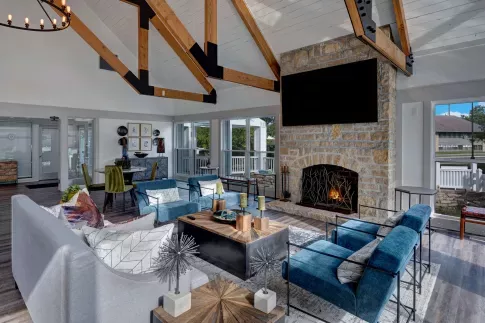Elegant resident lounge featuring vaulted ceilings with exposed wood beams, a floor-to-ceiling stone fireplace, plush modern seating, and abundant natural light from surrounding windows.
