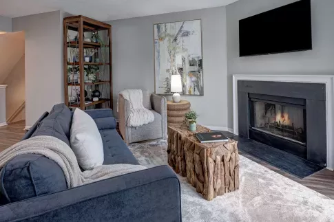 Cozy living room featuring a navy upholstered sofa, rustic wood coffee tables, a modern abstract painting, and a sleek black fireplace with a mounted TV above.