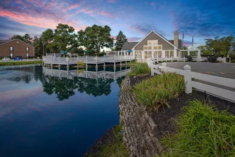 Peaceful twilight view of a lakeside clubhouse with a spacious white deck extending over calm water, surrounded by greenery, fencing, and beautifully reflected sky colors for a serene and picturesque atmosphere.