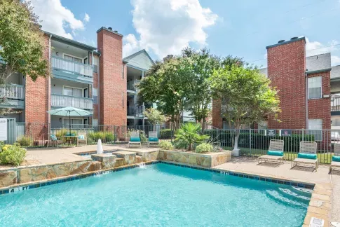 Sparkling swimming pool with a water feature and lounge chairs surrounded by trees and landscaping, with red brick apartment buildings and balconies in the background.