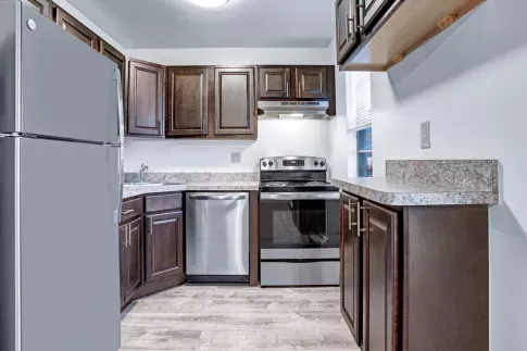 A contemporary kitchen at Laurel Run Village Apartments featuring dark wood cabinetry with brushed nickel hardware, stainless steel appliances including a refrigerator, dishwasher, and oven, and speckled granite countertops. The space is well-lit with a ceiling-mounted light fixture, and a small window adds natural light. The kitchen layout offers ample counter space with an extended bar area for additional functionality.