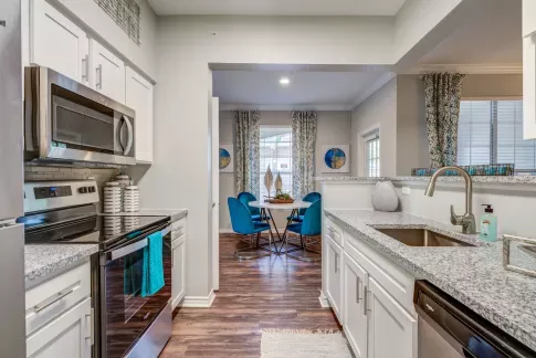 Modern galley kitchen with granite countertops and stainless steel appliances, opening to a dining area with blue velvet chairs and patterned drapes.