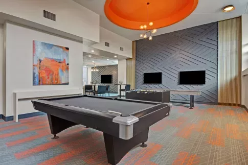 Modern resident lounge featuring a pool table, foosball, stylish geometric accent wall with mounted TVs, and vibrant contemporary décor under an eye-catching orange ceiling dome.