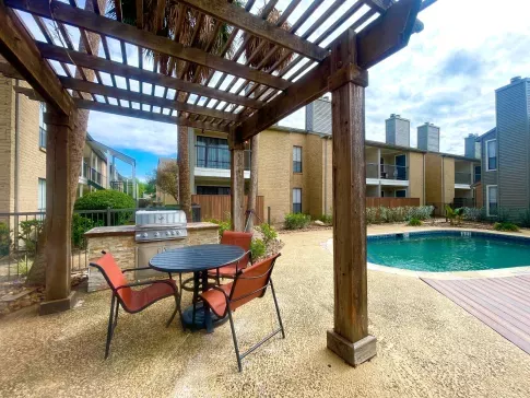 An inviting outdoor amenity space at Oaks of Westchase featuring a shaded pergola with a round dining table and four red chairs. Adjacent to the seating area is a built-in stainless steel grill for outdoor cooking. The area overlooks a sparkling swimming pool, surrounded by lush landscaping and modern apartment buildings with private balconies. The scene is framed by tall palm trees, creating a relaxed and resort-like atmosphere.