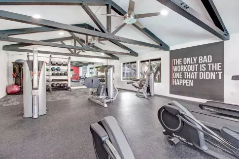 A modern fitness center at Olive Ridge Apartments, featuring high ceilings with exposed dark beams and large windows for natural light. The gym is equipped with state-of-the-art strength and cardio machines, free weights, and functional training accessories. A motivational wall mural reads, 'The only bad workout is the one that didn’t happen.'