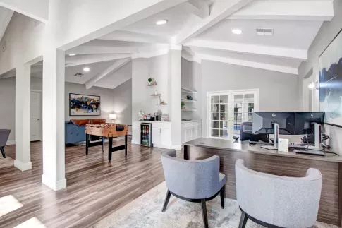 A stylish clubhouse and leasing office at Olive Ridge Apartments, designed with vaulted ceilings and open-concept seating areas. The space includes a foosball table, a cozy lounge area with a blue couch, and a reception desk with dual monitors. Decorative shelving, modern artwork, and natural light contribute to the inviting ambiance. ​​