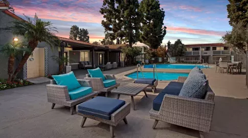 A resort-style swimming pool area at Olive Ridge Apartments captured during twilight. The space features woven wicker lounge chairs with plush blue and turquoise cushions, a communal seating area with ottomans, and a shimmering pool surrounded by lush landscaping. The evening sky is painted in shades of pink and blue, creating a serene atmosphere.