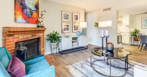 A stylish and contemporary living room at Santana Ridge featuring a cozy brick fireplace with vibrant abstract artwork above it. The room is furnished with a teal accent chair, a modern white console table adorned with decorative pieces, and a sleek glass coffee table with candles. The open-concept design leads to a dining area and kitchen with white cabinetry, creating a bright and inviting space. Warm wood flooring and indoor plants add a touch of comfort and elegance.