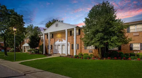 A grand entrance to Silver Lake Manor Apartments at twilight, showcasing a stately red brick building with tall white columns and an elegant gabled roof. The manicured lawn and vibrant flower beds add to the curb appeal, while a lamppost lights up the scenic sidewalk leading to the entrance. The sunset sky in the background creates a warm, inviting ambiance. ​​