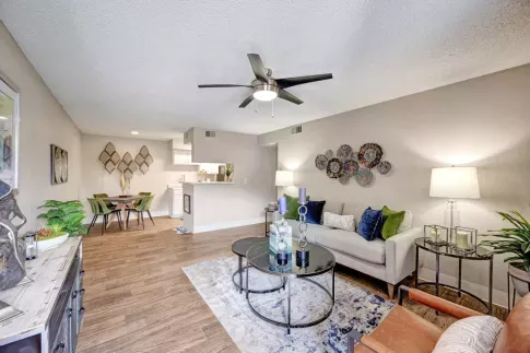 A modern open-concept living space at Skyline Apartments featuring a stylish living room, dining area, and kitchen. The living room is furnished with a gray sofa adorned with blue and green accent pillows, a round glass coffee table, and a leather accent chair. Decorative wall art, table lamps, and lush indoor plants enhance the ambiance. The dining area includes a wooden table with four green velvet chairs, while the kitchen features white cabinetry and a pass-through counter, creating a seamless and invit