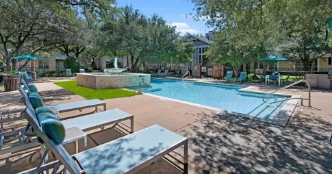A luxurious outdoor pool area in a residential community, surrounded by mature trees and lush landscaping. The pool features a built-in water fountain, lounge chairs with blue and green striped pillows, and patio tables with teal umbrellas. A modern clubhouse with large windows is visible in the background, adding to the relaxing, resort-like atmosphere. ​​