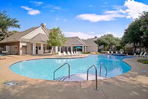 Resort-style swimming pool surrounded by lounge chairs and trees, with a view of the clubhouse and residential buildings in the background under a clear blue sky