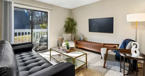 Modern living room at The Forest Apartments featuring a black leather sofa, a glass coffee table with a gold frame, and a cozy armchair with a throw blanket. A large sliding glass door leads to a private balcony with outdoor seating, allowing natural light to fill the space. The room is decorated with neutral tones, a sleek media console, and stylish accents, creating a warm and inviting atmosphere.