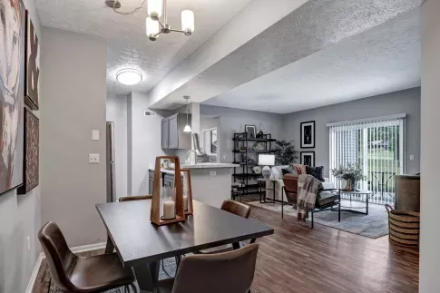A modern open-concept living space at Trotters Landing featuring a dining area, kitchen, and living room. The dining area has a dark wood table with four leather chairs and decorative candle lanterns. The kitchen includes gray cabinetry, stainless steel appliances, and a white island with pendant lighting. The living room is furnished with a cozy sofa, accent chairs, and a stylish bookshelf. Large sliding glass doors lead to a balcony with a green lawn view, allowing natural light to fill the space. ​​