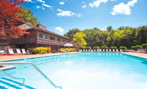 Outdoor swimming pool with clear blue water, surrounded by lounge chairs and umbrellas, next to a rustic two-story clubhouse with trees in the background on a sunny day.