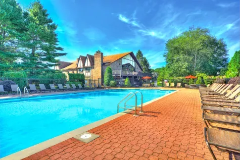 Bright outdoor swimming pool with rows of lounge chairs on a brick patio, surrounded by trees and a Tudor-style clubhouse with balconies and red umbrellas in the background.